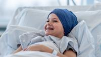 A young boy battling Cancer lays in his hospital bed tucked under the blankets. He is wearing a head scarf to keep him warm and has an optimistic smile on his face.